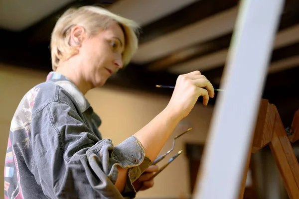 Wanita bahagia melukis di atas kanvas menggunakan kuas tipis. Kanvas berdiri di sel. Seniman menggambar di easel. Tampilan samping lukisan perempuan gambar di kanvas di studio rumah seni — Stok Foto