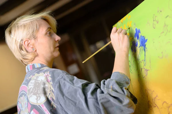 Donna felice dipinge su tela con pennello sottile. Tela sta sul cavalletto. Artista disegna al cavalletto. Vista laterale della pittura femminile quadro su tela in studio d'arte casa — Foto Stock