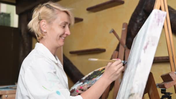 幸せな女性は薄いブラシを使用してキャンバス上でペイント。帆布はウナギの上に立っている。画家はイーゼルを描く。ホームスタジオにおけるキャンバス上の女性の絵画の側面図 — ストック動画