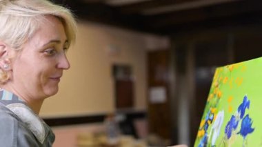 Mutlu kadın ince fırça kullanarak tuvale resim yapıyor. Tablo sehpanın üzerinde duruyor. Ressam sehpada resim yapıyor. Resim stüdyosunda tuvale resim çizen bir kadının yan görüntüsü