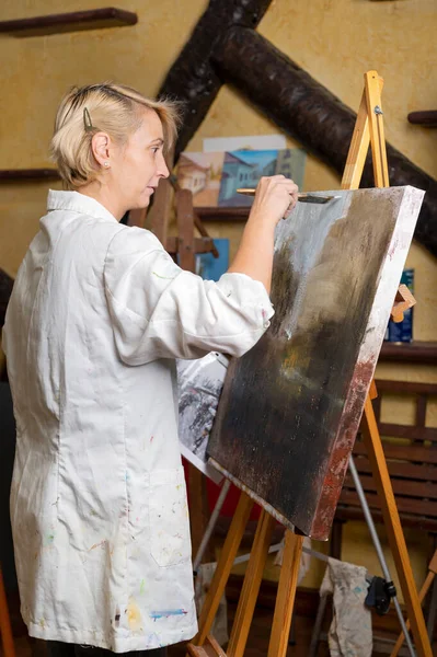 Donna felice dipinge su tela con pennello sottile. Tela sta sul cavalletto. Artista disegna al cavalletto. Vista laterale della pittura femminile quadro su tela in studio d'arte casa — Foto Stock