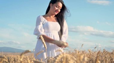 Güneşli bir günde altın buğday tarlasında dikilen beyaz elbiseli genç kadın buğday başaklarına hafifçe dokunuyor..