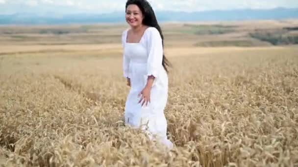 Ung latinamerikansk kvinna i vit klänning går mitt i ett vetefält och ler mot kameran. — Stockvideo