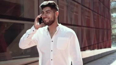 Genç, neşeli, pozitif adam dışarıda, ofis binalarının yakınında telefonla konuşurken yürüyor.. 