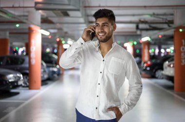 Arabasını park ettikten sonra telefonla konuşurken otoparkta yürüyen kendinden emin, genç ve yakışıklı bir adam..