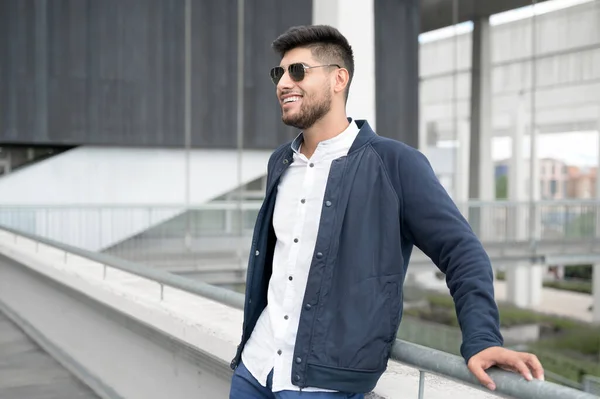 Портрет молодого уверенного улыбающегося человека, стоящего рядом с современным зданием и смотрящего вдаль — стоковое фото