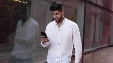 Sakallı adam telefon ekranına bakıyor ve sokakta dikilirken gülüyor. Akıllı telefon kullanarak sosyal medya haberlerini tarayan yakışıklı bir adam..