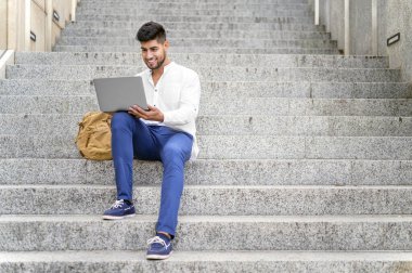 Merdivenlerde dizüstü bilgisayarla çalışan yakışıklı genç adam.