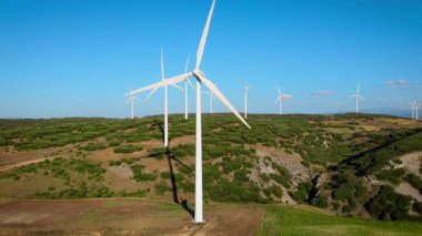Geniş rüzgar türbinlerinin açık mavi bir gökyüzünün altında yeşil tarlaların üzerinde döndüğü bir rüzgar çiftliğinin eril drone görüntüleri. Video, kırsal alanda sürdürülebilir teknoloji ve temiz enerji içeren yenilenebilir enerji üretimini yakalıyor..