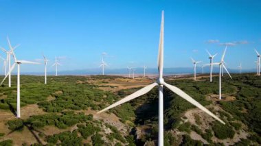 Geniş rüzgar türbinlerinin açık mavi bir gökyüzünün altında yeşil tarlaların üzerinde döndüğü bir rüzgar çiftliğinin eril drone görüntüleri. Video, kırsal alanda sürdürülebilir teknoloji ve temiz enerji içeren yenilenebilir enerji üretimini yakalıyor..