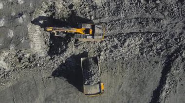 Açık maden ocağındaki bir çöp kamyonuna taş yükleyen bir kazıcının yukarıdan aşağı görüntüsü. Endüstriyel İHA görüntüleri, ağır makinelerin tozlu bir ortamda çalıştığını gösteriyor. Çıkartma, madencilik ve inşaat faaliyetlerini vurguluyor. Yüksek kalite 4k 