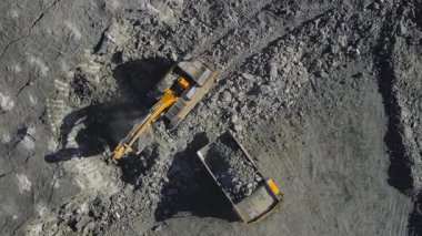 Açık maden ocağındaki bir çöp kamyonuna taş yükleyen bir kazıcının yukarıdan aşağı görüntüsü. Endüstriyel İHA görüntüleri, ağır makinelerin tozlu bir ortamda çalıştığını gösteriyor. Çıkartma, madencilik ve inşaat faaliyetlerini vurguluyor. Yüksek kalite 4k 