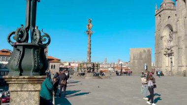 Porto, Portekiz 'deki Katedral Meydanı' nda güneşli bir gün. İnsanlar taştan meydanda yürüyorlar. Etrafı tarihi binalarla çevrili. Ortaçağ kulesi ve süslemeli sütun gibi parlak mavi bir gökyüzünün altında. Yüksek kalite 4k görüntü