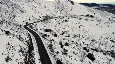 Karlı bir kış manzarasının içinden geçen dolambaçlı bir dağ yolunu izleyen hava aracı videosu. Kavisli asfalt, beyaz tepeler, uzak doğa, soğuk hava, sinematik seyahat manzarası.