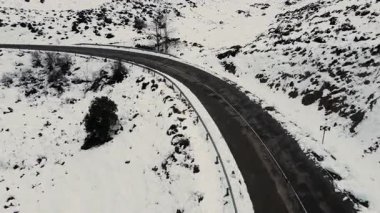 Karlı bir kış manzarasının içinden geçen dolambaçlı bir dağ yolunu izleyen hava aracı videosu. Kavisli asfalt, beyaz tepeler, uzak doğa, soğuk hava, sinematik seyahat manzarası.