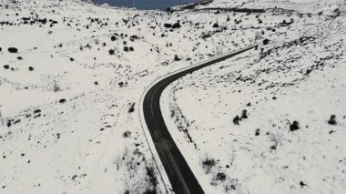 Karlı bir kış manzarasının içinden geçen dolambaçlı bir dağ yolunu izleyen hava aracı videosu. Kavisli asfalt, beyaz tepeler, uzak doğa, soğuk hava, sinematik seyahat manzarası.