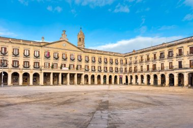 Vitoria-Gasteiz 'deki İspanya meydanının geniş bir görüntüsünde neoklasik cepheler, balkonlar ve parlak mavi bir gündüz vakti gökyüzünün altında açık bir taş meydan yer alıyor. Yüksek kalite fotoğraf