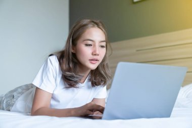 Güzel bir Asyalı kadın uzanmış, yatağa ve dizüstü bilgisayarına gülümsüyor. WFH karantinada evden çalışıyor. Sosyal Mesafe Öz İzolasyonu