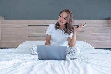 Yatağa uzanmış, dizüstü bilgisayarı ile gülümseyen bir kadın. O, iş hakkında konuşmak için telefonda. WFH karantinada evden çalışıyor. Sosyal Mesafe Öz İzolasyonu