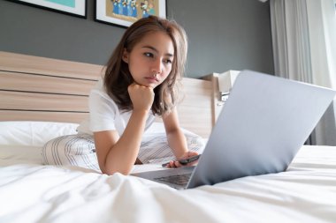 Güzel bir Asyalı kadın uzanmış, yatağa ve dizüstü bilgisayarına gülümsüyor. WFH karantinada evden çalışıyor. Sosyal Mesafe Öz İzolasyonu