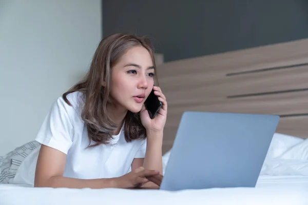 Smiling woman lying down the bed and her laptop. She is on the phone to talk about work. WFH Working from home in quarantine lockdown. Social distancing Self Isolation