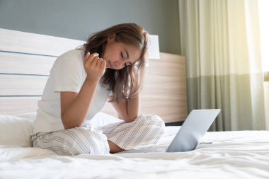 Güzel bir Asyalı kadın uzanmış, yatağa ve dizüstü bilgisayarına gülümsüyor. WFH karantinada evden çalışıyor. Sosyal Mesafe Öz İzolasyonu