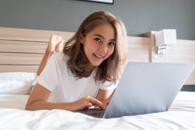 Güzel bir Asyalı kadın uzanmış, yatağa ve dizüstü bilgisayarına gülümsüyor. WFH karantinada evden çalışıyor. Sosyal Mesafe Öz İzolasyonu