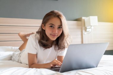 Güzel bir Asyalı kadın uzanmış, yatağa ve dizüstü bilgisayarına gülümsüyor. WFH karantinada evden çalışıyor. Sosyal Mesafe Öz İzolasyonu