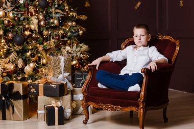 Sekiz yaşında beyaz gömlekli bir çocuk Noel ağacının yanındaki sandalyede oturuyor.