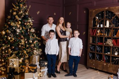Beş kişilik güzel, mutlu bir aile Noel ağacının yanında duruyor ve kameranın içine bakıyor. Anne, baba ve üç çocuk.