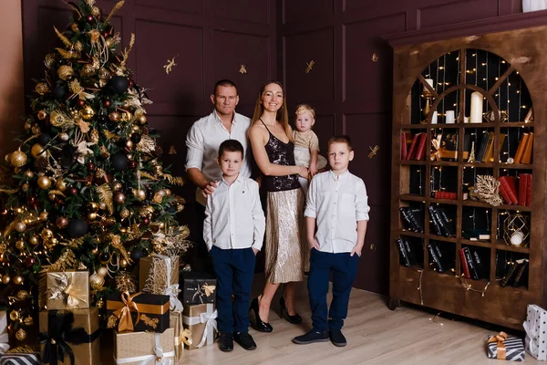 A beautiful happy young family of five stands in full height near the Christmas tree and look into the camera indoors. mother, father and three children