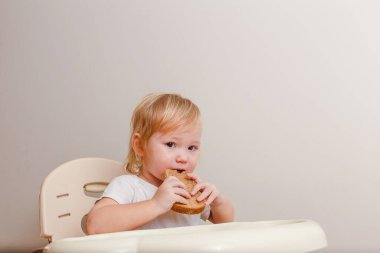Beyaz elbiseli şirin bir buçuk yaşında bir kız beslenme koltuğunda oturup beyaz arka planda ekmek yiyor.