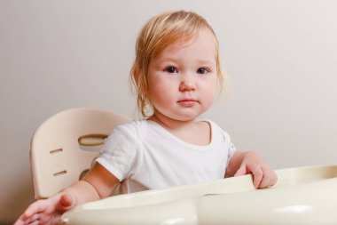 Beyaz elbiseli tatlı sarışın küçük kız beyaz arka planda bebek koltuğunda oturuyor.