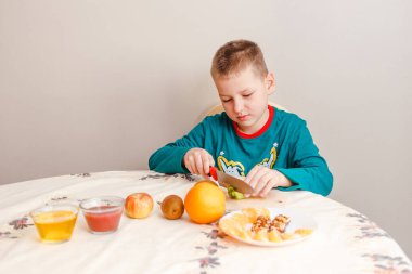 Dokuz yaşında bir çocuk masada oturur ve kivi doğrar. Çocuk evde kendi kahvaltısını hazırlar.