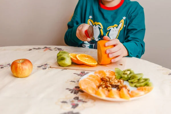 9 yaşındaki bir çocuk bir masada oturur ve bir portakal keser. Çocuk evde kendi kahvaltısını hazırlar.