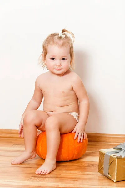 A one-and-a-half-year-old blonde girl in panties sits on a large orange pumpkin on a white background