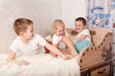 Üç neşeli çocuk yatakta oynuyor. Çocuklar evde çıplak ayakla birbirlerini gıdıklıyorlar..