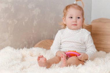 Bir buçuk yaşında sarışın bir kız evdeki yatakta oturan bir oyuncakla oynuyor.