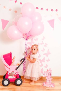 Güzel pembe elbiseli mutlu sarışın kız evde oyuncak bebek arabasıyla oynuyor. Bir yıllık fotoğraf çekimi