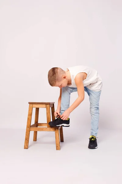 Nine-year-old European boy puts his foot on a stool and ties his shoelaces on a white background