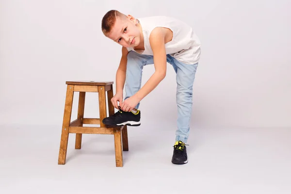 Nine-year-old European boy puts his foot on a stool and ties his shoelaces on a white background