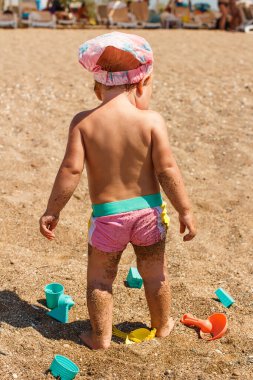 Bir yaşında bronzlaşmış bir kız çocuğu yazın kumsalda sırtı dönük durur.