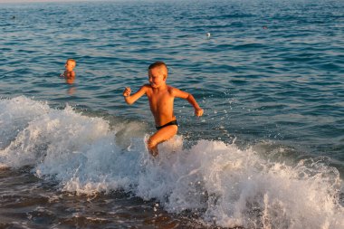 Bronzlaşmış bir çocuk gün batımında denizde suyla koşar.