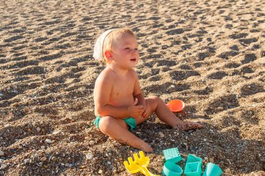 Mayolu şirin bir kız kumsalda kumsalda oturur ve spatulayla oynar. Yazın bronzlaşmış bebek