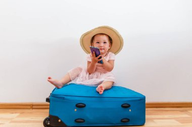 Hasır şapkalı, elinde telefonla içeride büyük bir bavula oturan sevimli bir kız.