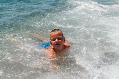 Bronz tenli, kısa saçlı dokuz yaşındaki çocuk deniz kenarında yüzüyor ve yazın dalgalarla oynuyor..