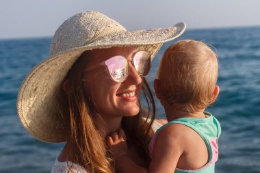 Şapkalı ve gözlüklü gülümseyen genç bir kadın bir yaşındaki bebeği kollarında denizde tutuyor.