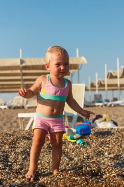 Pembe mayo giyen bir yaşındaki sarışın kız Türkiye 'de kumlu bir çakıl taşı plajında çıplak ayakla duruyor.