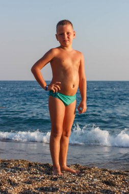 İyi beslenmiş, mayolu gülümseyen bir çocuk denizin kenarında duruyor.