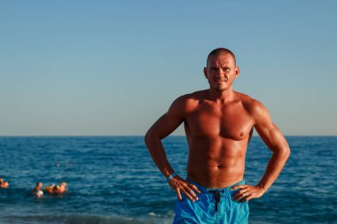 Şişirilmiş kaslı mavi şortlu vahşi adam denize karşı duruyor.